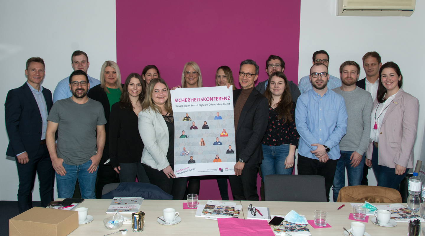 Teilnehmer der Sicherheitskoferenz stehen gemeinsam mit Plakat vor magenta-Wand